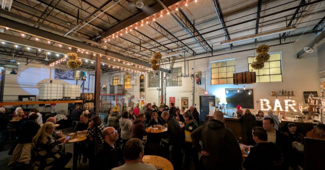 A happy group of people sitting at a long bar and around tables, enjoying an event in a distillery A happy group of people sitting at a long bar and around tables, enjoying an event in a distillery