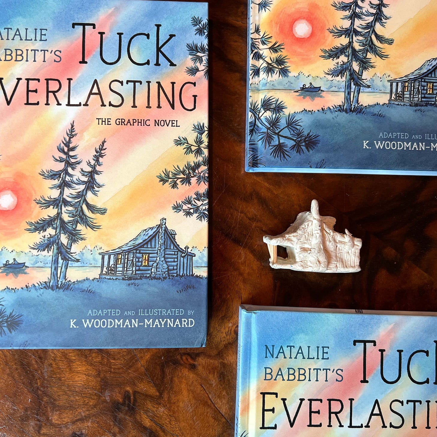 Cover image of K. Woodman-Maynard's graphic novel adaptation of Tuck Everlasting with a reference clay house model beside it.