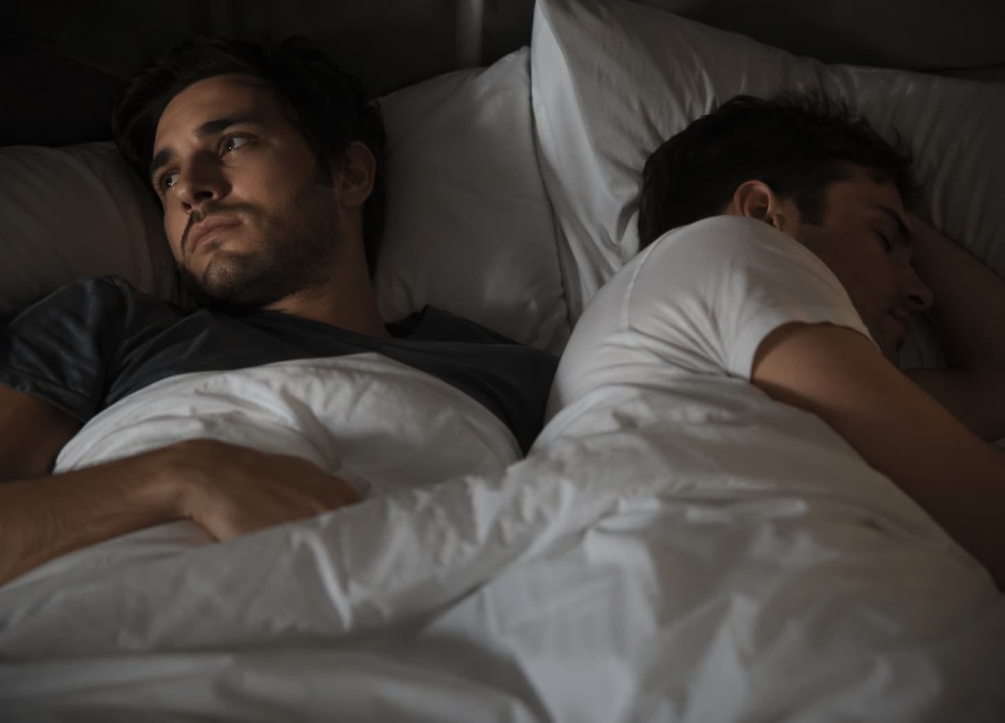 A man lies awake in a dimly lit bedroom, staring off with a tense, distant expression, while his partner sleeps peacefully beside him under white sheets, turned away and unaware.