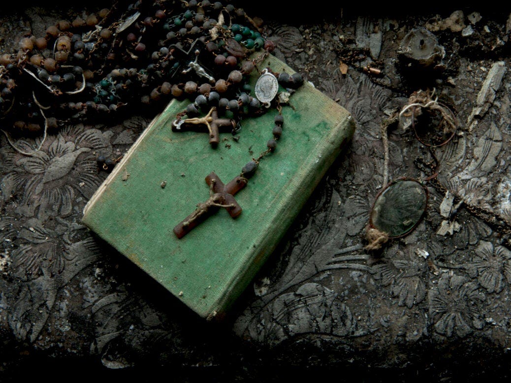 Photo shows a crucifix at the end of a rosary, lying on green book with candle and broken spectacles against wallpaper.