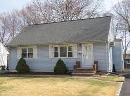 Light blue clapboard single-story house with gray shingled roof and white trim on windows and door. Front yard features patchy grass, two small evergreen shrubs flanking the concrete steps to the white front door. Bare trees and early spring sky in background. Attached garage on right side. Street visible with yellow lines. Light blue clapboard single-story house with gray shingled roof and white trim on windows and door. Front yard features patchy grass, two small evergreen shrubs flanking the concrete steps to the white front door. Bare trees and early spring sky in background. Attached garage on right side. Street visible with yellow lines.