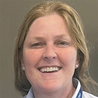 Retrato em close de uma mulher adulta que sorri para a câmera. Ela tem cabelos loiros claros, presos para trás, e usa camisa clara com pequenos padrões, além de um crachá com cordão azul no pescoço. Retrato em close de uma mulher adulta que sorri para a câmera. Ela tem cabelos loiros claros, presos para trás, e usa camisa clara com pequenos padrões, além de um crachá com cordão azul no pescoço.