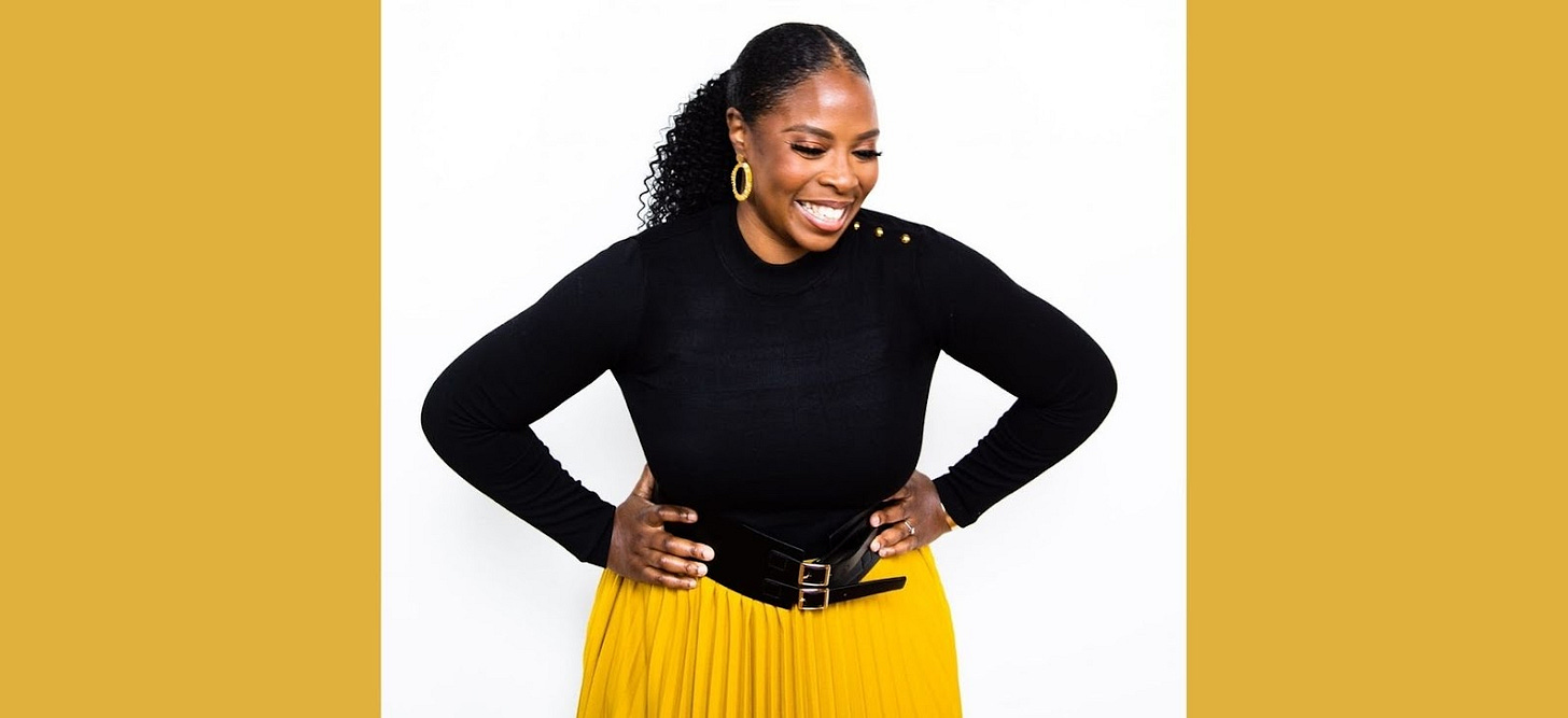 Photo of Stephanie with her hand on her hips, smiling and looking downward, wearing a black long sleeved top and a bright yellow skirt.