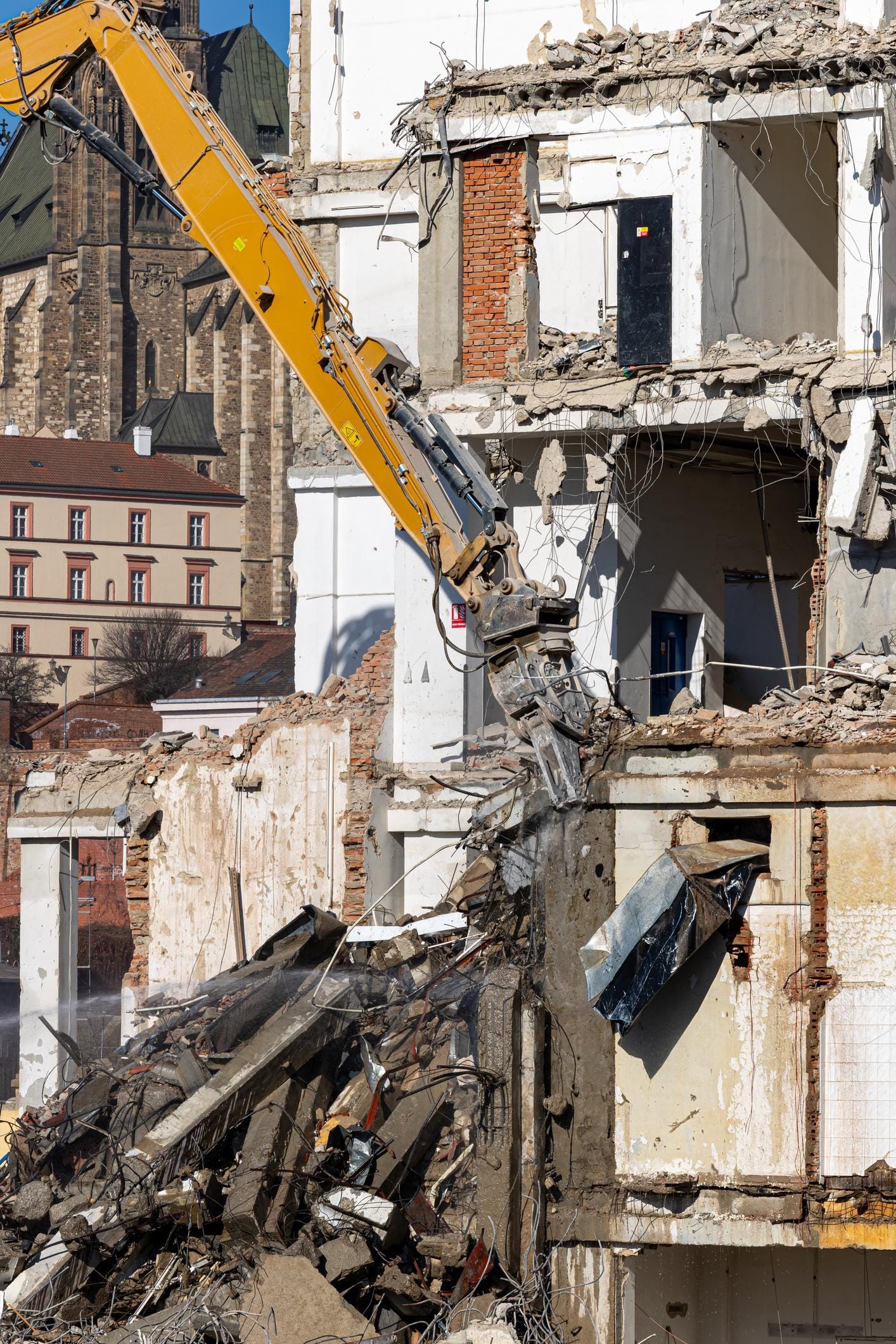 Urban demolition in progress with a long-reach excavator tearing apart a damaged concrete building, piles of rubble below, and a historic cathedral rising in the background Urban demolition in progress with a long-reach excavator tearing apart a damaged concrete building, piles of rubble below, and a historic cathedral rising in the background