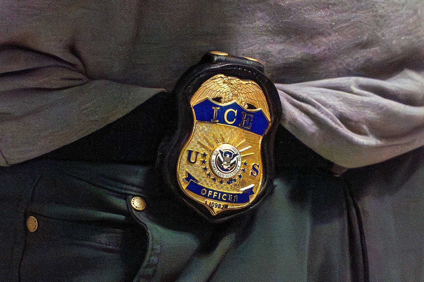 An ICE badge is on the waist of someone wearing a very sloppily tucked-in gray athletic tee and baggy black pants.
