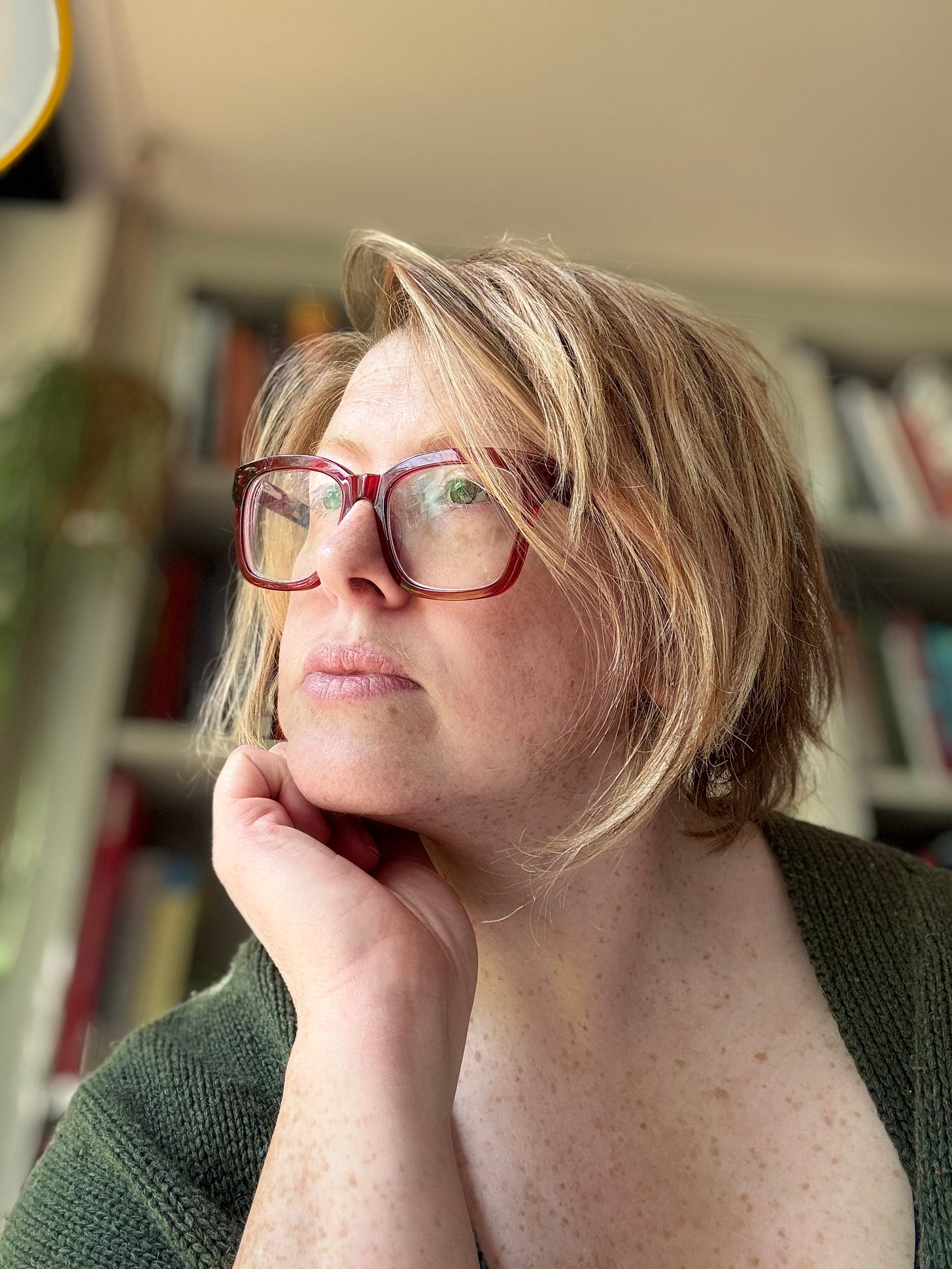 Jenny, a light-skinned person with reddish-blonde hair cut in a shaggy bob and red glasses, looks out away from the camera with their chin resting on their hand. 