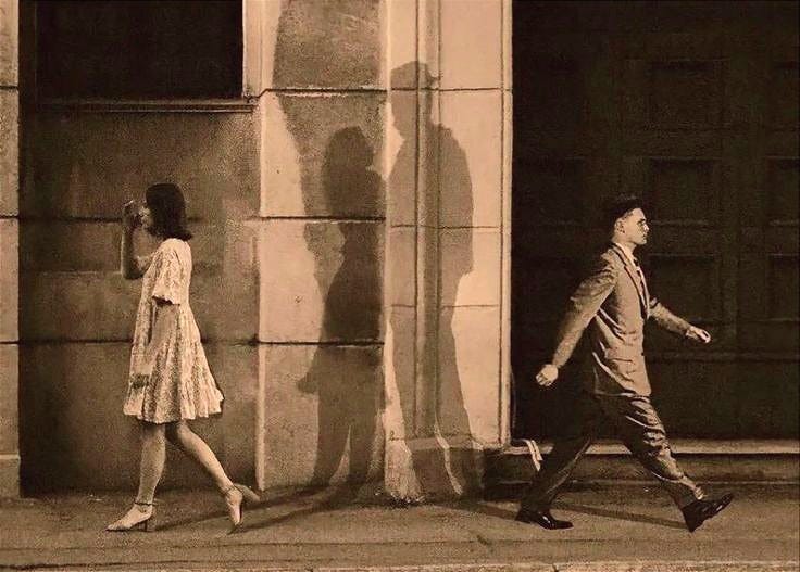 This may contain: a man and woman walking down the street in front of a building with shadows on it