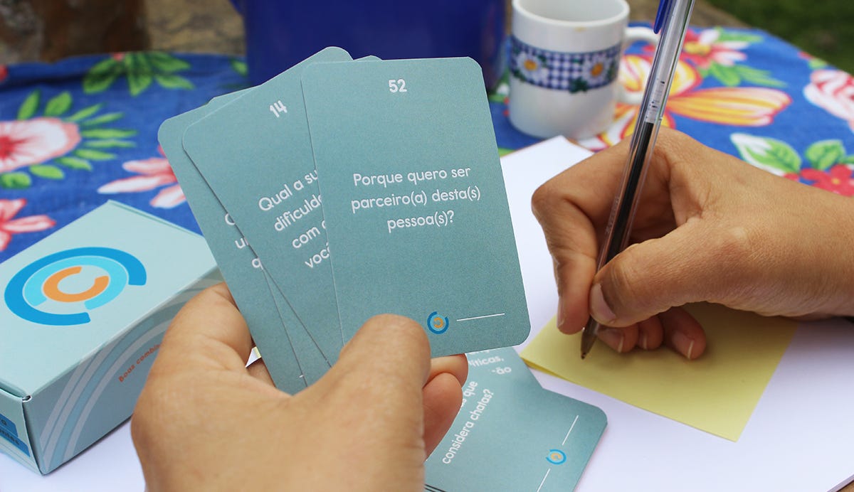 duas mãos aparecem na foto, uma segurando algumas cartas, onde na primeira se lê "Porque quero ser parceiro(a) desta(s) pessoa(s)?" e a outra segura uma caneta para anotar as reflexões. as mãos estão em cima de uma mesa, com toalha de chita azul, ao fundo estão uma xícara de café e uma jarra azul, além da caixinha do jogo Combinados, que é cinza e tem o logo formato por vários Cs, um dentro de outro, nas cores azul, azul claro e laranja.