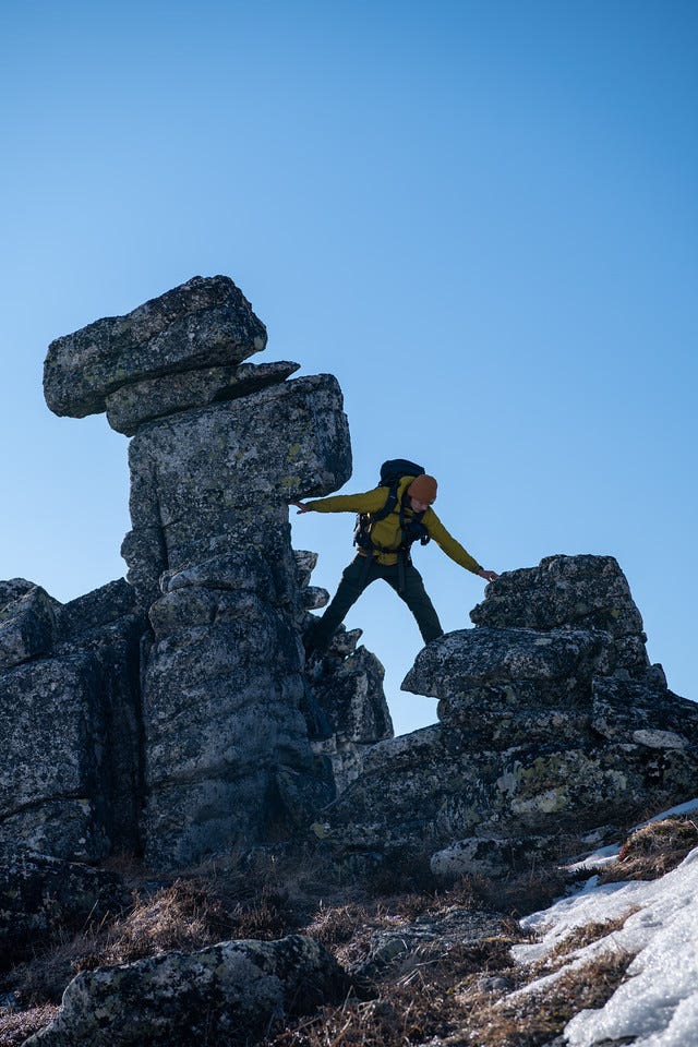 A hiker in a yellow top and pack steps across a gap between weathered granite blocks, one stack capped by a balanced boulder, against a clear blue sky.