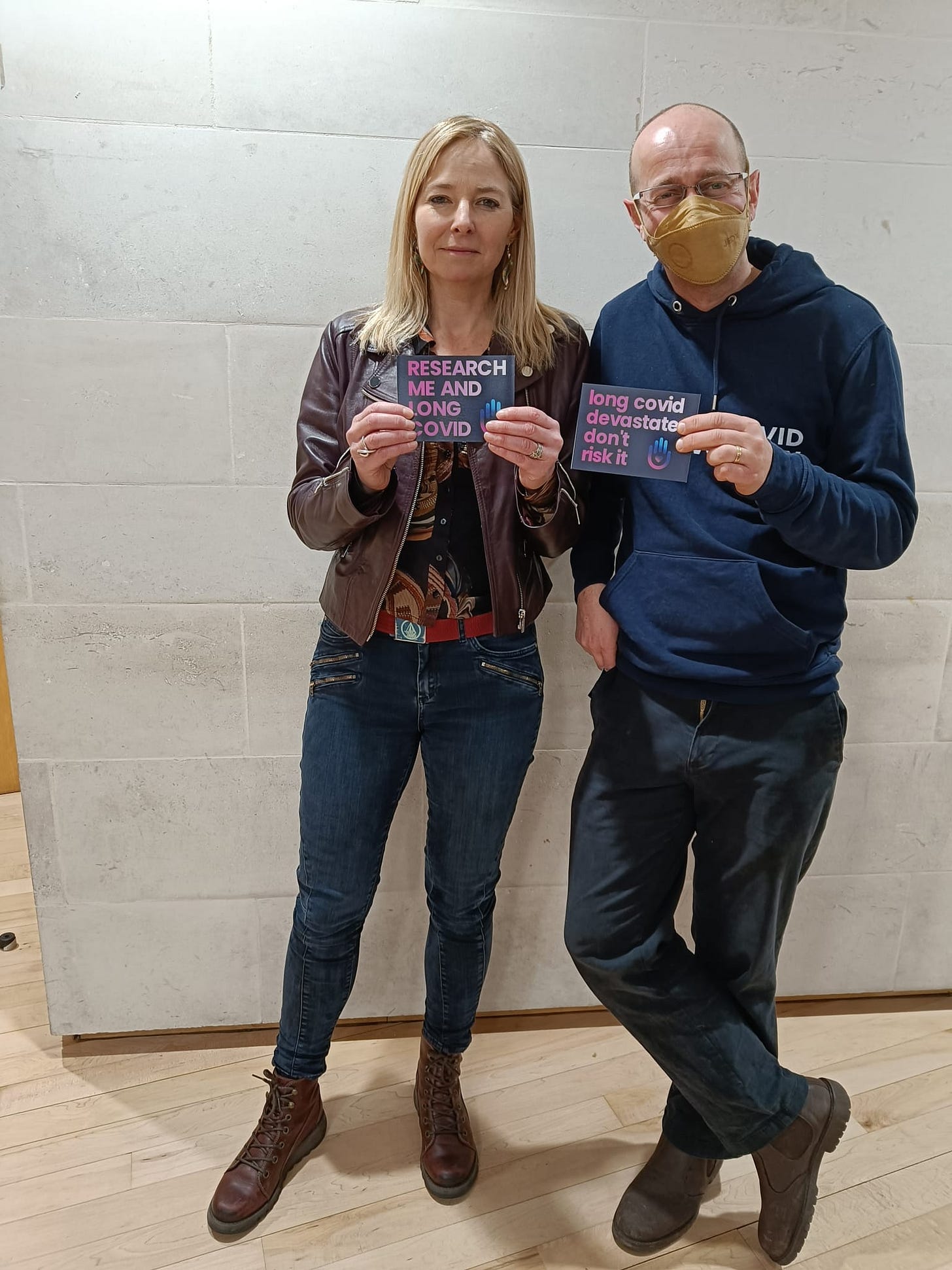Two people hold signs about long COVID awareness against a white wall. One wears a mask. Text reads "Research me and Long COVID" and "Long COVID devastates, don't risk it."