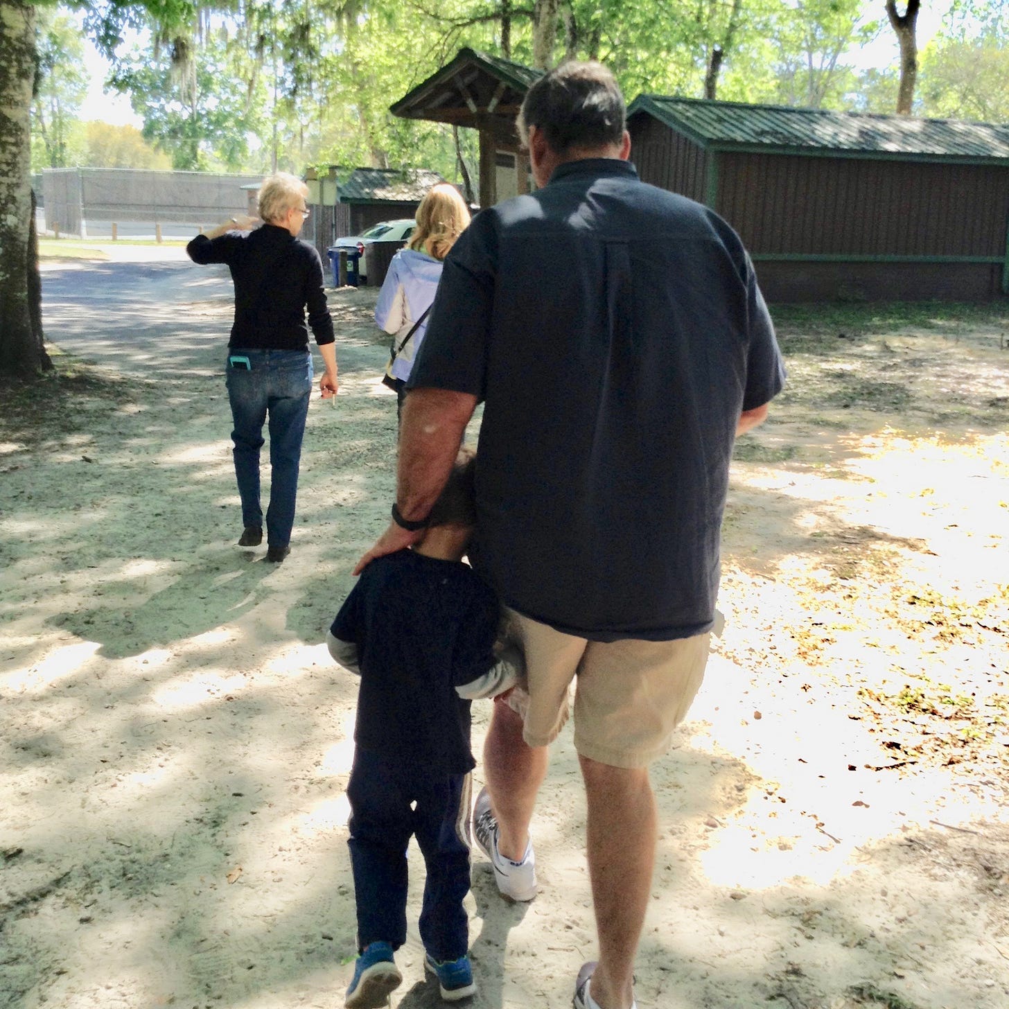 Well-built man in a navy short sleeved shirt and khaki shorts walking in a clearing with a small boy.  The man’s head is slightly bent and has his hand on the left shoulder of the boy.  The boy has his arm wrapped around the man’s left leg, with his head nestled in between the man’s left hand and the man’s left hip.