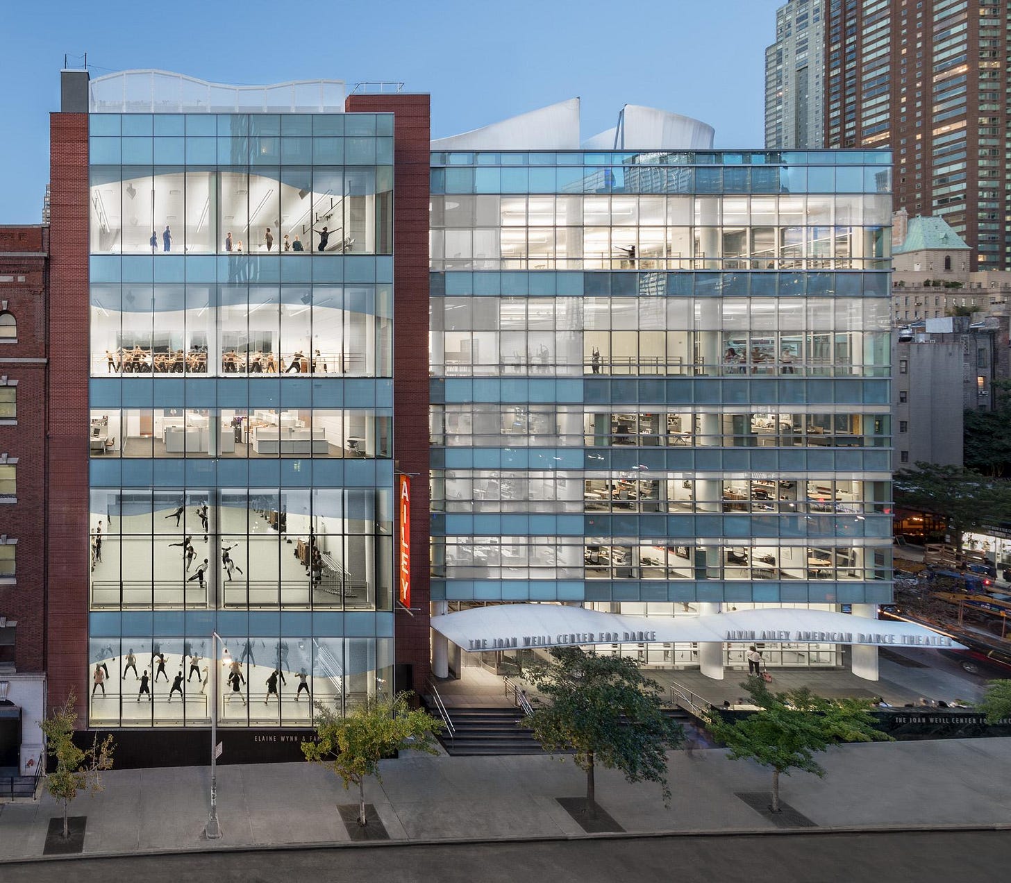 Exterior view of The Joan Weill Center for Dance with the Elaine Wynn & Family Education Wing. The modern building has large glass windows, showing multiple floors with people practicing dance inside. The building is surrounded by other tall buildings in an urban setting.
