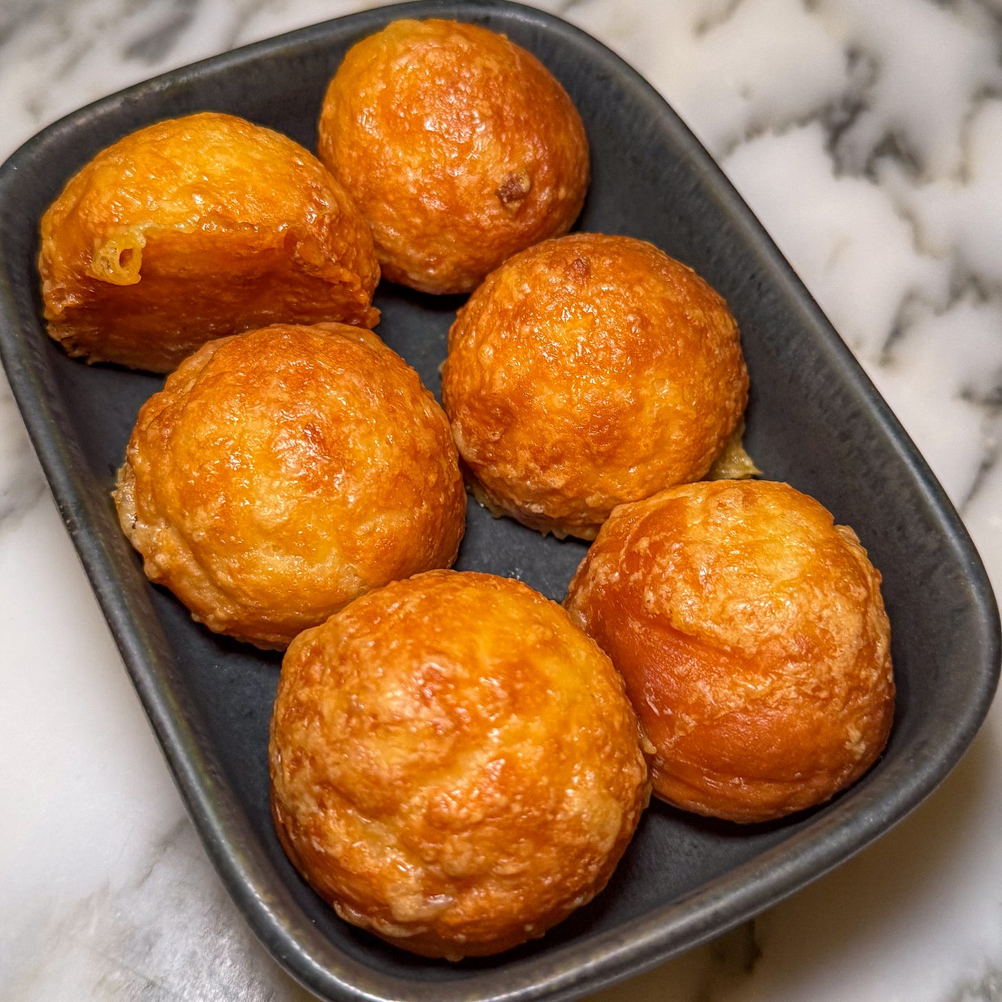 Six golden, round Comté gougères served warm in a small dark dish