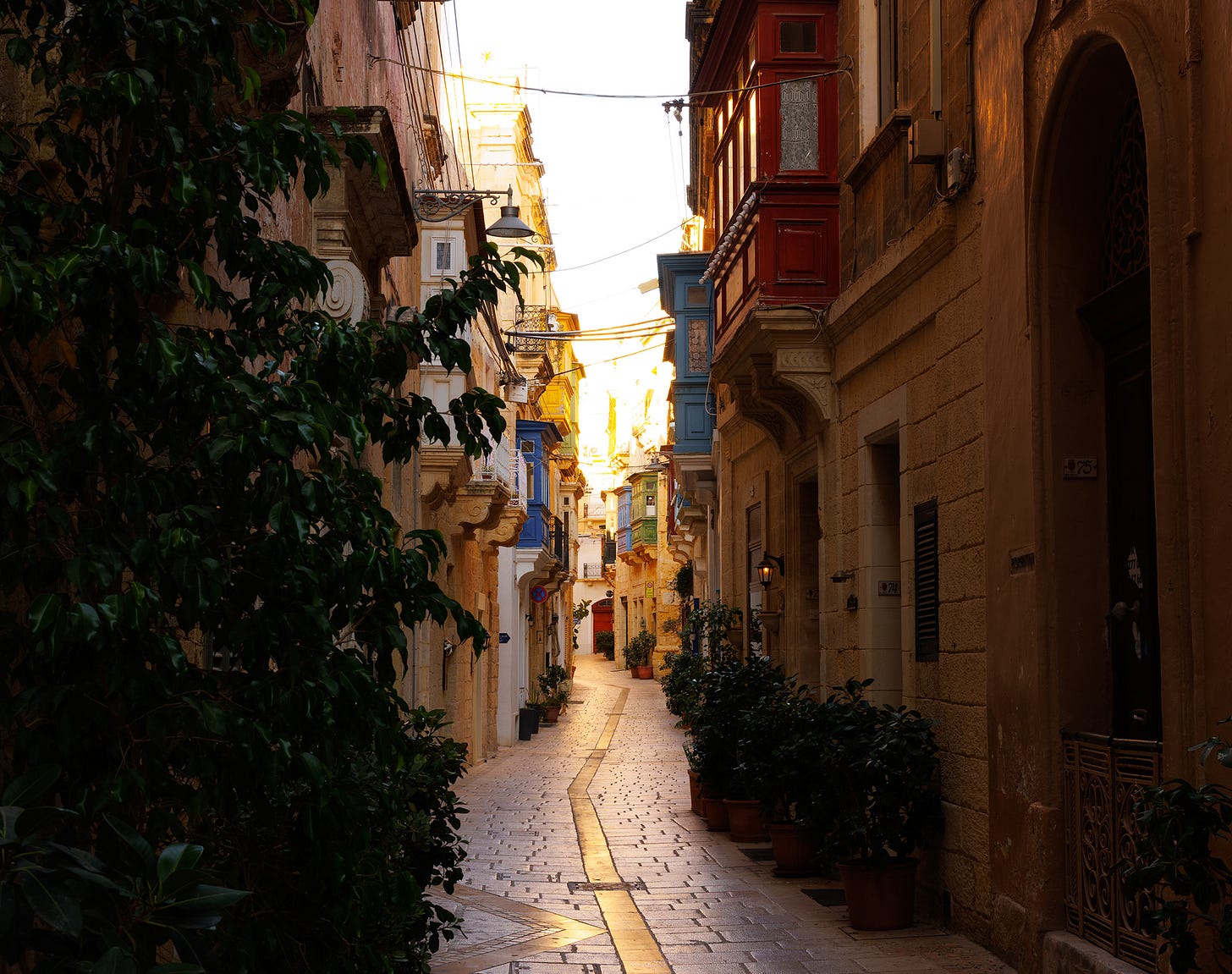 Triq Tabone Street in Birgu, Malta, a narrow stone alley paved with worn limestone blocks. The passage curves gently toward the light, where the golden evening sun illuminates traditional Maltese balconies painted in red and blue. Potted plants line the lower walls, lanterns hang from the facades, and the close stone buildings press in, giving the street an intimate, lived-in character. This was once the nerve center of Christendom during the Great Siege, yet today it feels like a quiet neighborhood thoroughfare.