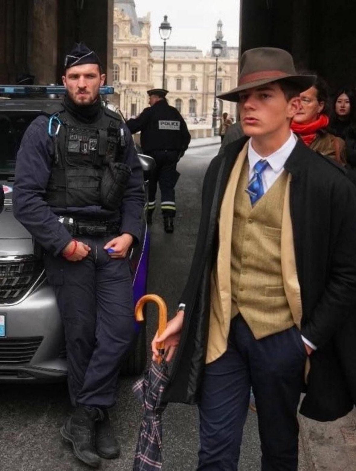 Man in tweed suit, tan vest, blue tie, black overcoat, fedora hat, and trousers holds purple umbrella and stands next to French police officer in black beret, tactical vest, and boots near gray police car outside grand arched entrance of Louvre museum with columns, lamps, and pedestrians in background including woman in red scarf.