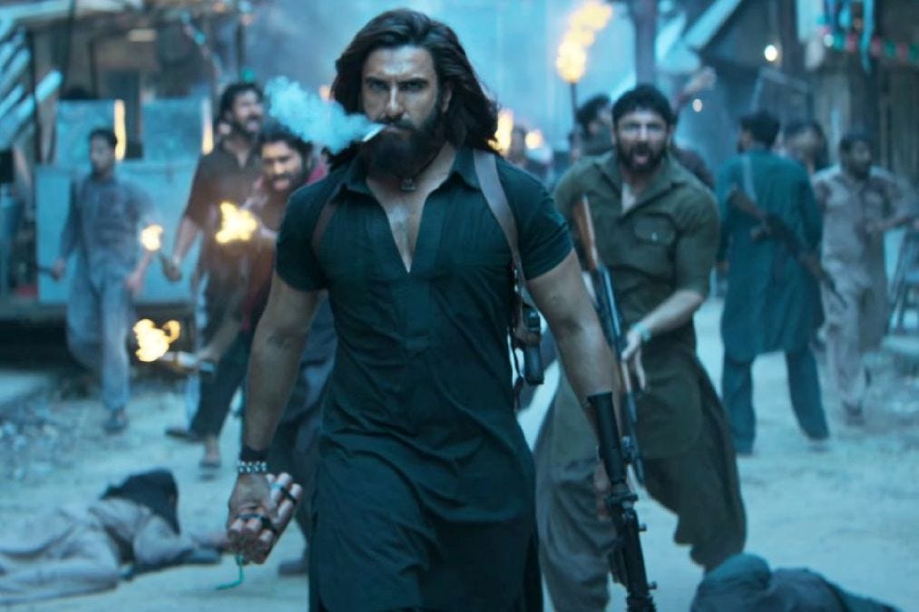 A still from Hindi film, ‘Dhurandhar (2025)’. Bearded protagonist Hamza (Ranveer Singh) strides down a narrow, battered street in a black kurta, smoke trailing from a cigarette in his mouth, a gun in one hand and explosives in the other. Behind him, armed men in dark clothes charge forward with rifles and flaming torches, while blurred figures and a fallen body lie in the background.