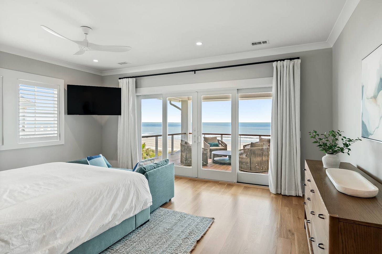A bedroom with a white-linen bed, white oak floors, a blue patterned rug, and sliding glass doors opening onto a porch with the ocean beyond. A bedroom with a white-linen bed, white oak floors, a blue patterned rug, and sliding glass doors opening onto a porch with the ocean beyond.