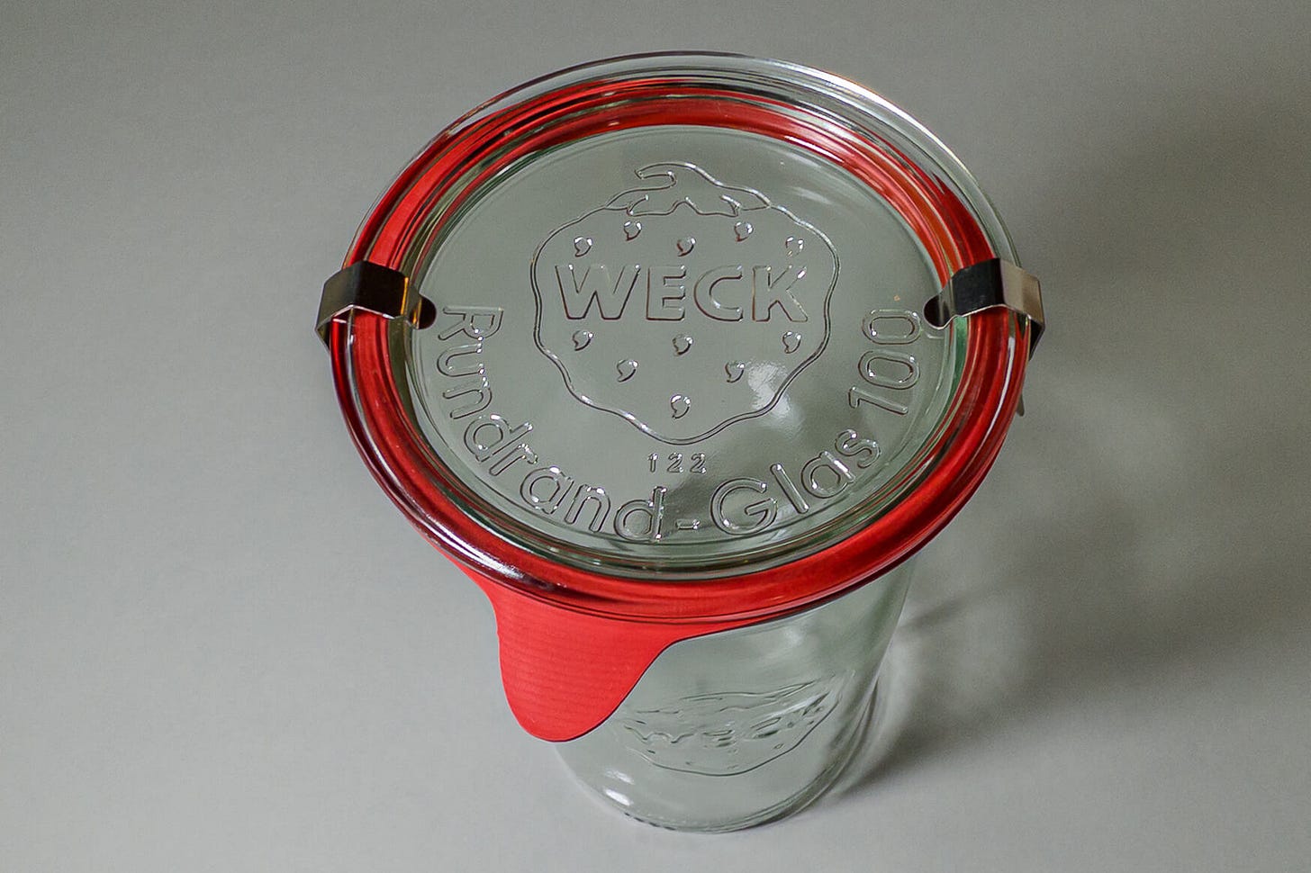 Top view of a sealed WECK round-rim jar with glass lid, orange rubber ring, and two metal clamps; the embossed WECK logo with its strawberry emblem and the words “Rundrand-Glas 100” are visible on the lid. Top view of a sealed WECK round-rim jar with glass lid, orange rubber ring, and two metal clamps; the embossed WECK logo with its strawberry emblem and the words “Rundrand-Glas 100” are visible on the lid.