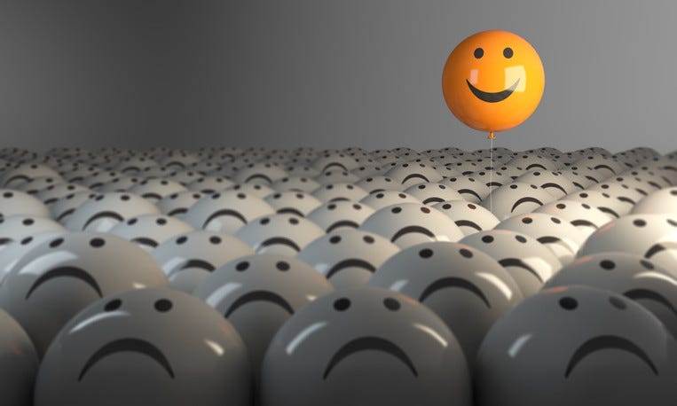 A field of glossy round black and white frown faces with one black and yellow smiing balloon rising above those