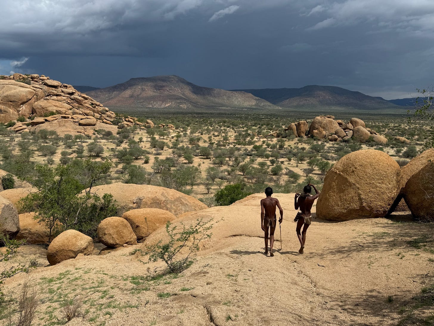 Two members of the San tribe walking across a rocky, arid landscape in Namibia under a dramatic, cloudy sky. Two members of the San tribe walking across a rocky, arid landscape in Namibia under a dramatic, cloudy sky.