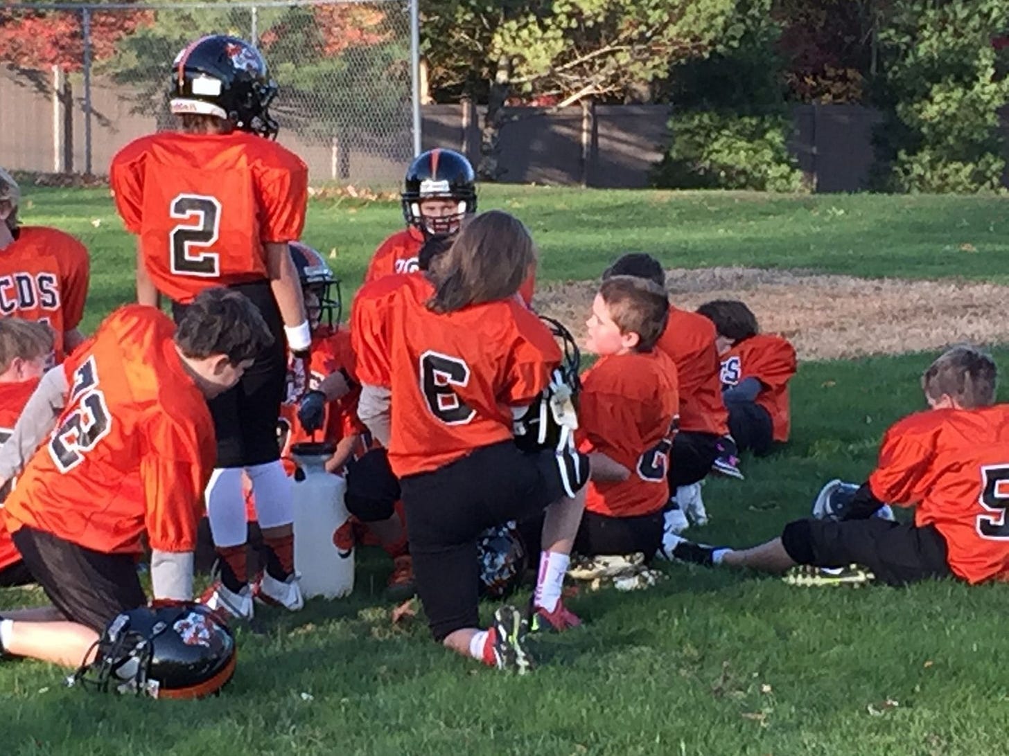 Myself, center, kneeling on a grass field surrounded by boys my age. We are wearing orange football uniforms and pads. Most of the boys have their helmets on while mine is off and my hair falls down the back of my jersey.