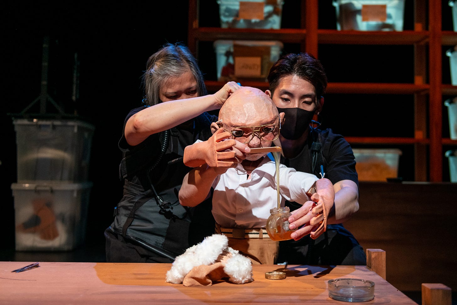 Two puppeteers dressed in black are holding a puppet. The puppet, a bestacled, bald old man, is holding a pot of glue. Two puppeteers dressed in black are holding a puppet. The puppet, a bestacled, bald old man, is holding a pot of glue.