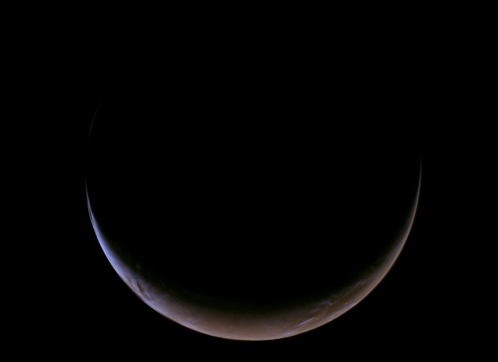 A slender, glowing crescent of Mars curves across the bottom of a completely black void, revealing its dusty reddish-orange surface and a thin, hazy blue atmospheric glow along the illuminated edge.