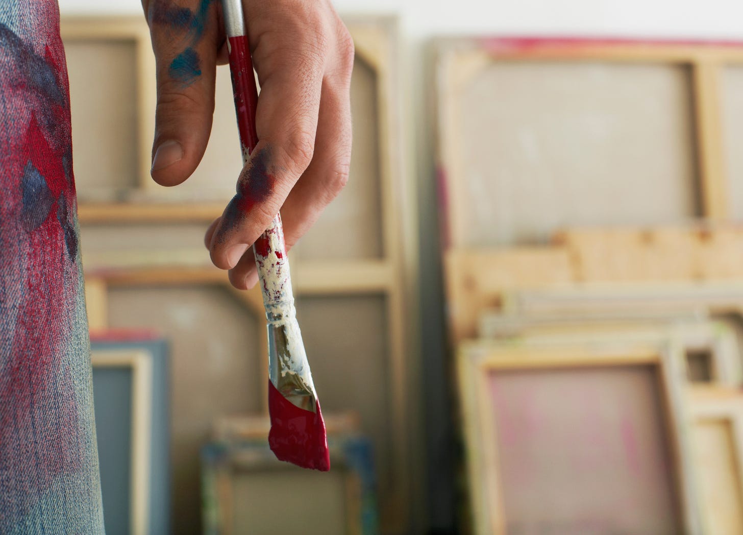 Close-up of an artist’s hand holding a paintbrush with red paint, wearing paint-streaked pants, with large blank canvases leaning against a wall in the background.
