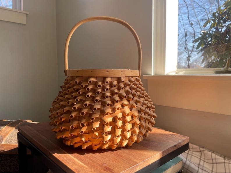 May include: A woven basket with a light brown color and a handle. The basket is made of thin strips of wood that are woven together to create a unique pattern. The basket is sitting on a wooden surface.