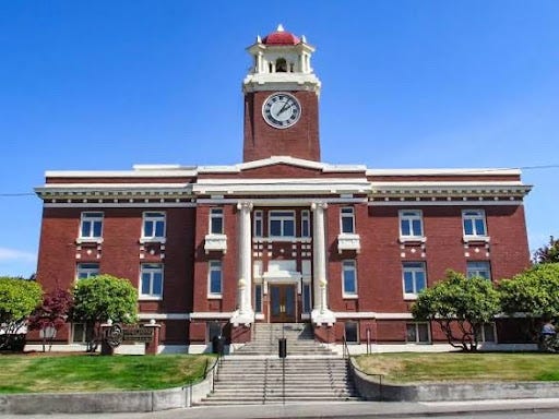A front-facing, professional photograph of the historic Clallam County Courthouse building in Port Angeles, emphasizing its large, multi-tiered concrete staircase, white classical columns, red brick facade, and central clock tower with a red-domed cupola under a clear blue sky.