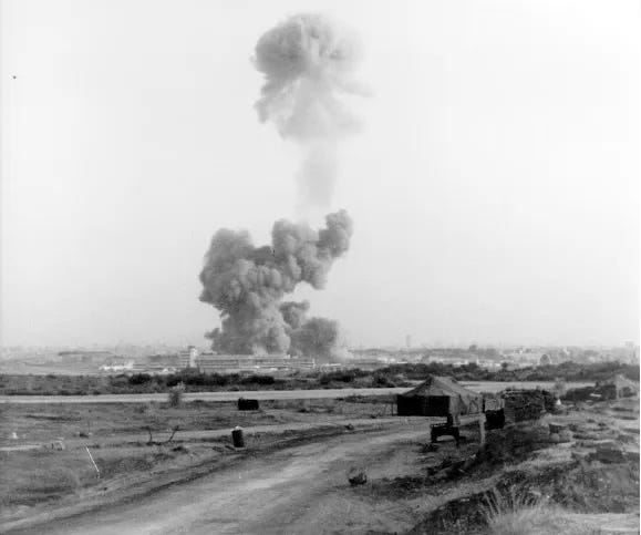 Black-and-white photograph of a massive explosion in the distance, with a towering column of smoke rising high above the skyline of Beirut. In the foreground, a dirt road curves past low sandbagged positions and small structures, while the city and blast site are visible across a flat, open landscape.