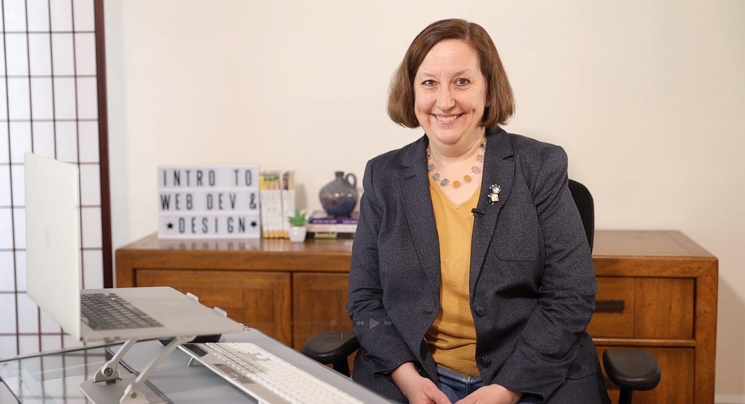 Jen Kramer sitting at a desk with computer and keyboard, a wooden chest behind her with books stacked on it. 
