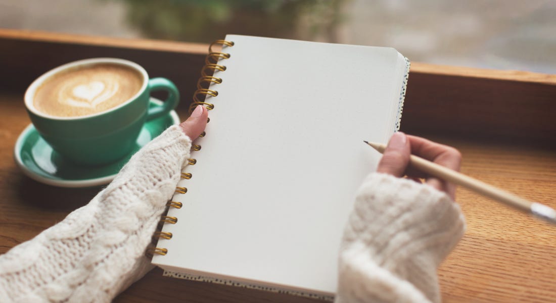 Two hands wrapped in a white cable-knit jumper holding a white spiral-bound notebook, with a blue coffee cup to the left of them and a desk behind the notepad Two hands wrapped in a white cable-knit jumper holding a white spiral-bound notebook, with a blue coffee cup to the left of them and a desk behind the notepad