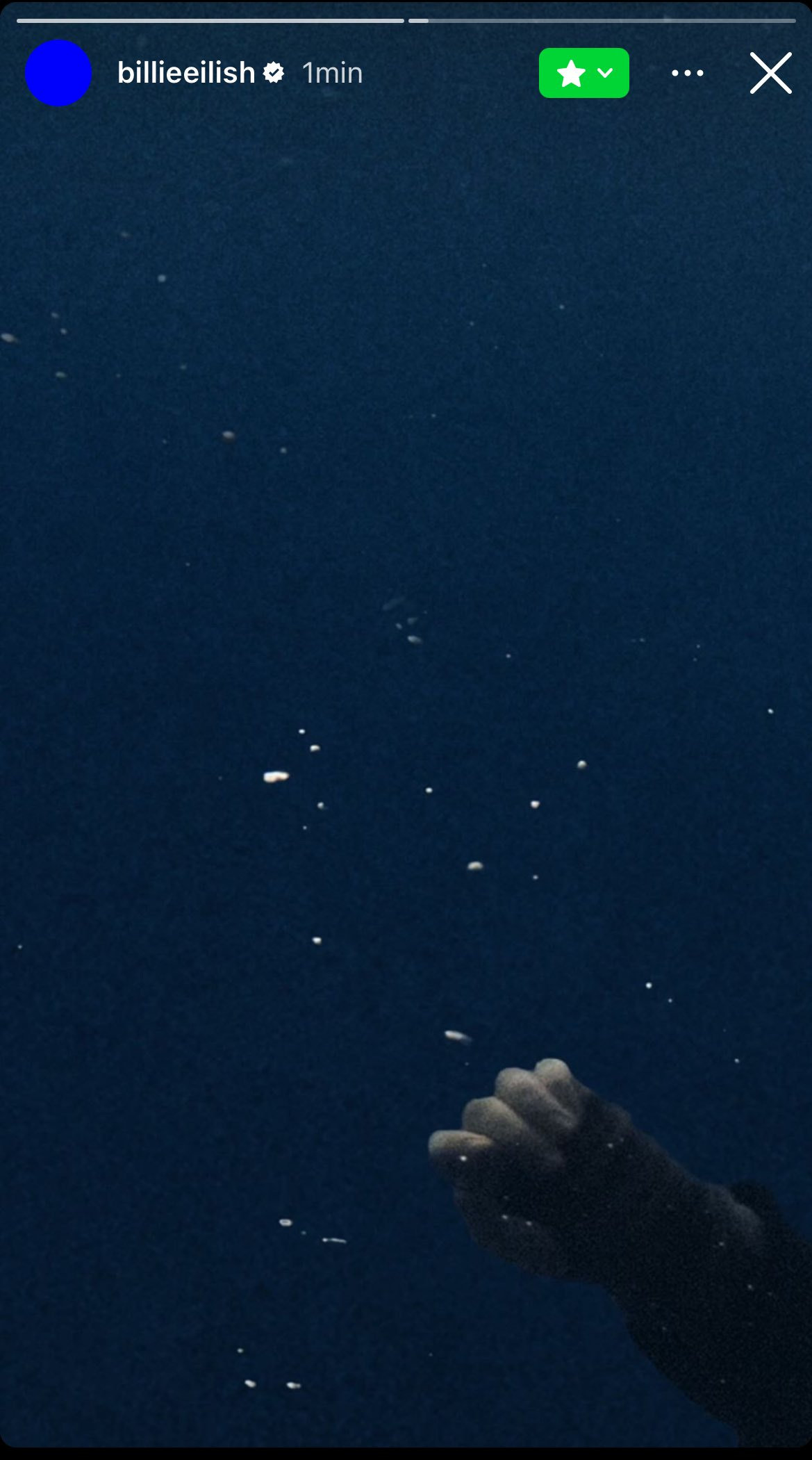 Printscreen de um story da cantora Billie Eilish, divulgado em seu close friends do Instagram. A imagem é predominantemente escura, com um fundo azul profundo que sugere água ou um ambiente submerso. No canto inferior direito, há uma mão parcialmente visível, fechada em um punho, com pequenas partículas brancas dispersas ao redor, como se fossem bolhas ou detritos flutuando.