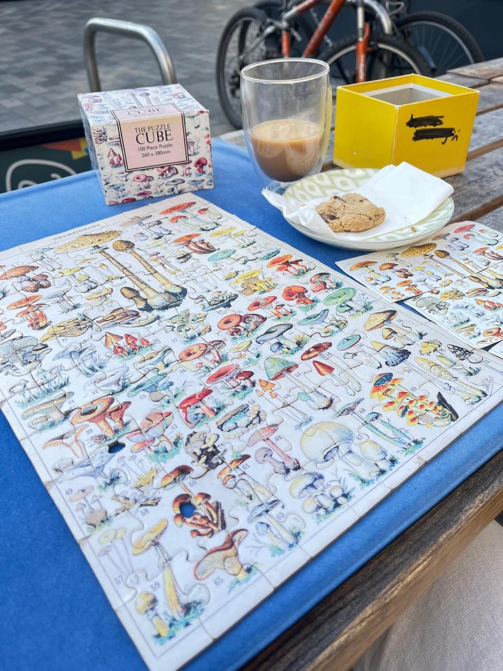 Image 1) Photo of a completed fungi botanical artwork style puzzle at a neurodivergent friendly cafe called Puzzle Bored in Brighton, UK. 2) Picture of Brighton beach on a sunny spring day with members of an event run by Leanne Maskell called 'AuADHD by the Sea'. The group holds up an artwork picture created by Leanne. Dog also in attendance