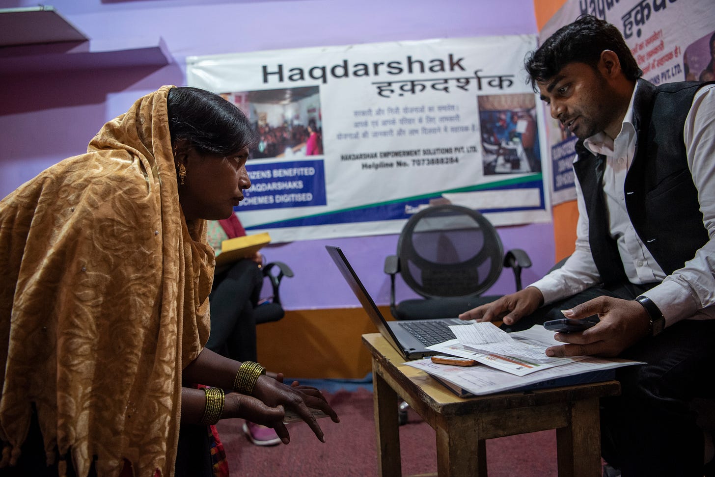An old woman having a conversation with a young man who's on his laptop. A haqdarshak banner is visible in the background