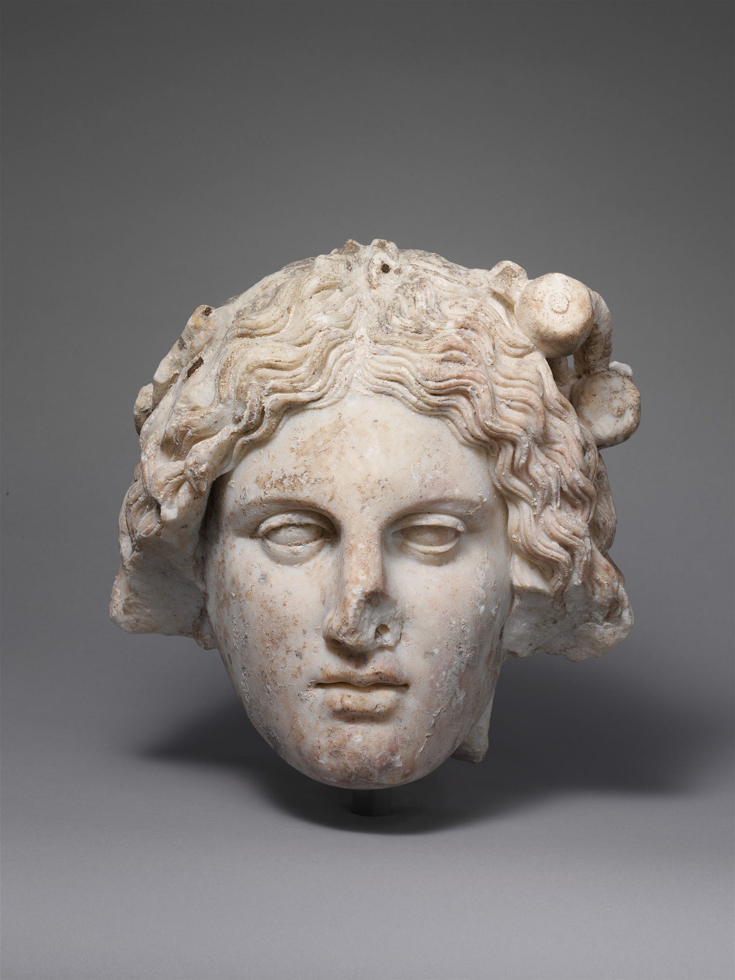 Photograph of the head from a Roman sculpture of a goddess, probably Ceres. The head is sculpted from marble. Her wavy hair is parted in the middle and is dressed so that it comes down just below her ears. She has an oval face with small, delicate lips. Her eyes have no pupils and she seems to be gazing ahead. Part of her nose is broken off, as is part of her hair. Pomegranate fruits woven into her hair suggest that she is a goddess associated with spring. The sculpture is in the collection of the Metropolitan Museum and dates to the first half of the 2nd century, CE. Photograph of the head from a Roman sculpture of a goddess, probably Ceres. The head is sculpted from marble. Her wavy hair is parted in the middle and is dressed so that it comes down just below her ears. She has an oval face with small, delicate lips. Her eyes have no pupils and she seems to be gazing ahead. Part of her nose is broken off, as is part of her hair. Pomegranate fruits woven into her hair suggest that she is a goddess associated with spring. The sculpture is in the collection of the Metropolitan Museum and dates to the first half of the 2nd century, CE.