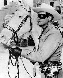 A masked cowboy in a gray outfit stands beside a white horse, holding the reins, with a western-style belt and holster, in a rustic desert setting.