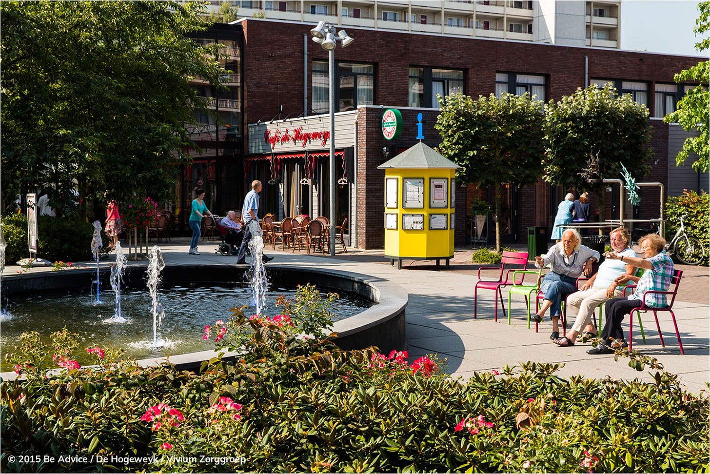the plaza at this dementia village features a wide fountain, mature trees and shrubs and chairs for sitting, surrounded by low-scale "commercia;" buildings and a two-story housing residence in the background the plaza at this dementia village features a wide fountain, mature trees and shrubs and chairs for sitting, surrounded by low-scale "commercia;" buildings and a two-story housing residence in the background