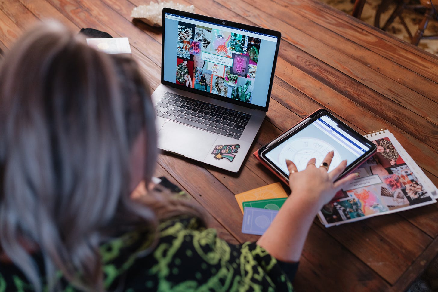Katie working at her laptop with a museboard pulled up and a birth chart and oracle cards on the table Katie working at her laptop with a museboard pulled up and a birth chart and oracle cards on the table