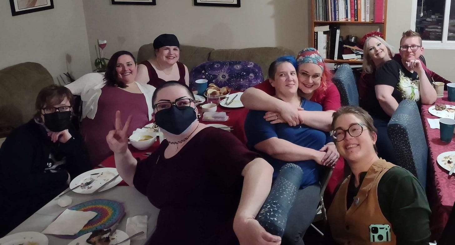 Nine queer people of all sorts gather around tables after a meal.