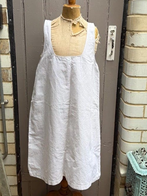 May include: A white linen dress with wide shoulder straps is displayed on a wooden dress form. The dress has a simple, classic design and hangs to about knee length. The background includes a wooden door, a ladder, and other vintage items.