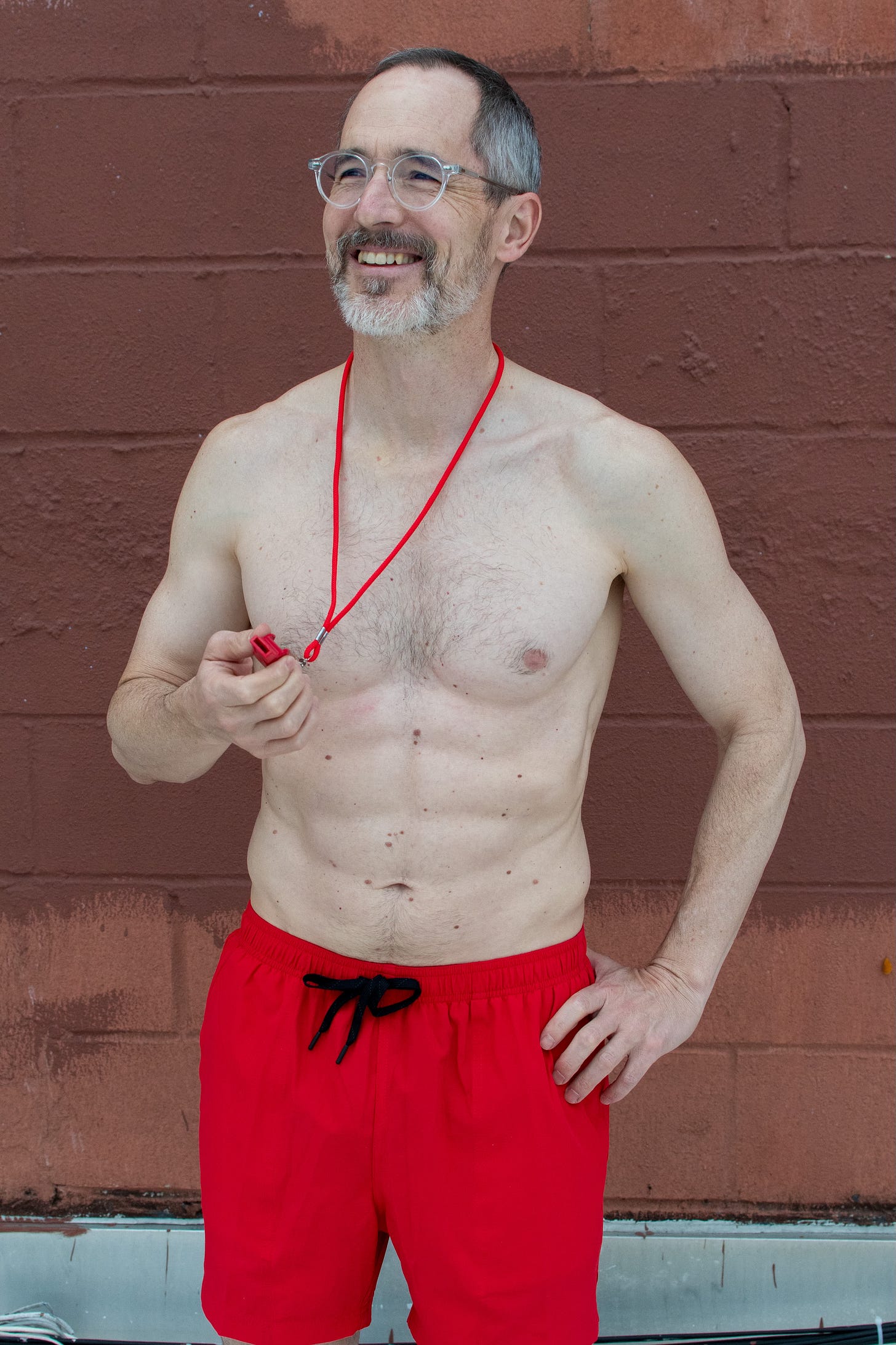 A shirtless middle-aged novelist in a bright red swimsuit, holding a bright red whistle, as he pretends it isn't late October. 