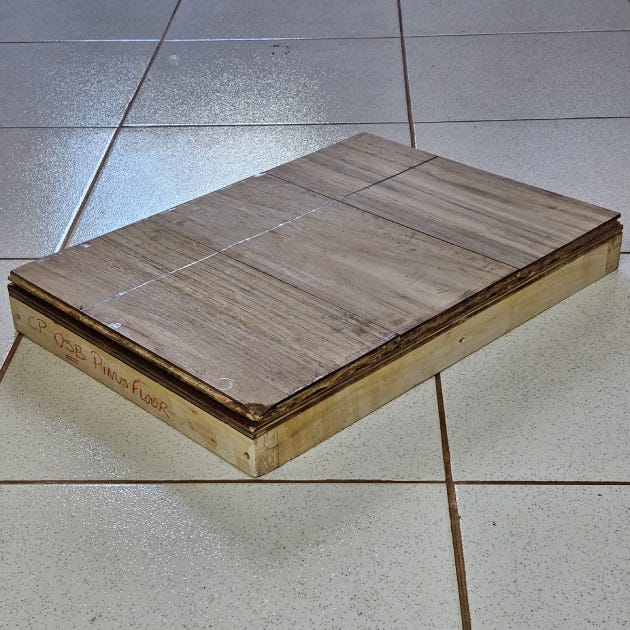 Fotografia de um painel estrutural de madeira repousando sobre um piso cerâmico. O painel é composto por múltiplas camadas de madeira prensada, com uma superfície que imita um acabamento de piso. A lateral do painel tem inscrições escritas à mão, sugerindo identificação para pesquisa ou testes laboratoriais. A iluminação natural reflete no piso, conferindo brilho ao ambiente.