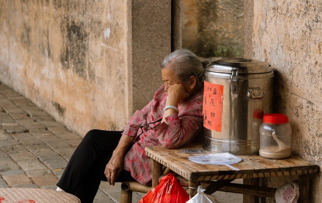 An elderly woman rests against a wall.