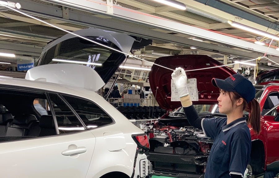 A Toyota factory worker stands beside a partially assembled vehicle, pulling the andon cord to pause production. The gesture reflects jidoka—automation with a human touch—where humans and machines collaborate to maintain quality and stop the line when issues arise.