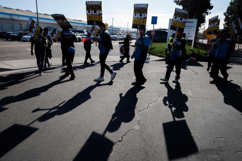 Photograph of protesters wearing blue amazon vests and holding signs marching in a picket line Photograph of protesters wearing blue amazon vests and holding signs marching in a picket line