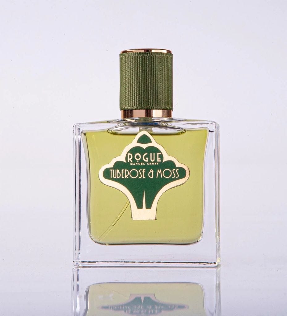 A glass bottle of Rogue Tuberos & Moss perfume featuring a green cap and label on a reflective surface.
