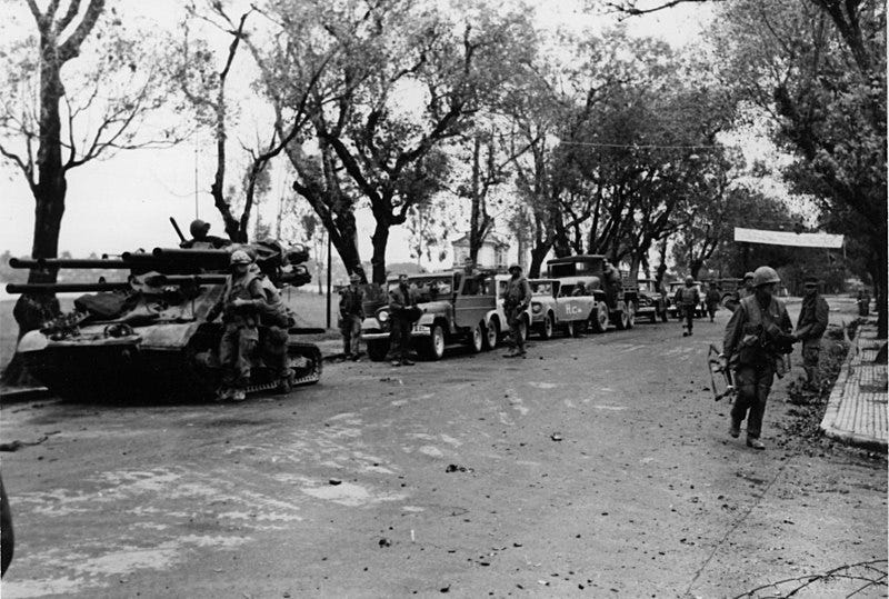 This black and white photo captures U.S. Marines and South Vietnamese forces during the Battle of Hue in the 1968 Tet Offensive.  Marines and soldiers move cautiously along a war-torn street, with an M50 Ontos armored vehicle and commandeered civilian trucks in the background. The battle was one of the longest and bloodiest of the Vietnam War, as U.S. and South Vietnamese troops fought to retake the city from North Vietnamese and Viet Cong forces in intense urban combat.