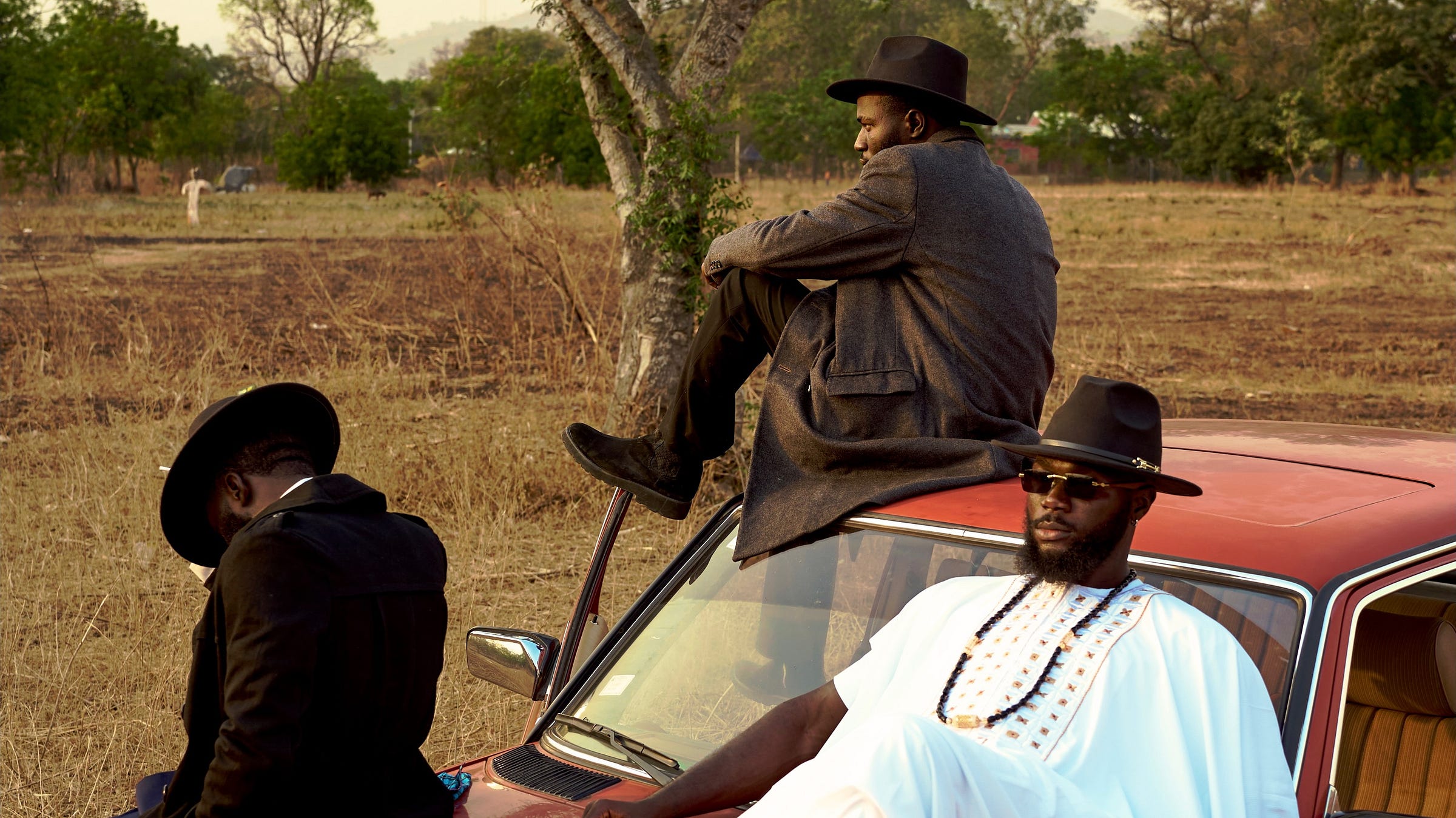 Three Nigerian men in hats posed on and around a vintage red Mercedes in golden-hour savanna